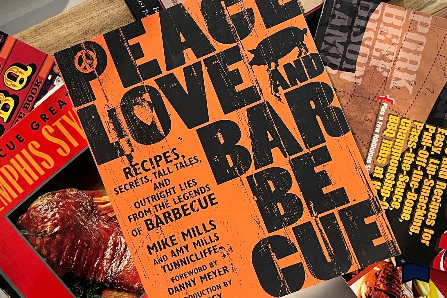 Collection of cookbooks on a wooden surface with 'Peace Love and Barbecue' prominently displayed.